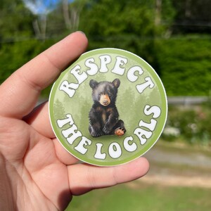 May include: A round sticker with a green background and white text that reads "Respect the Locals". A black bear cub is sitting in the centre of the sticker.
