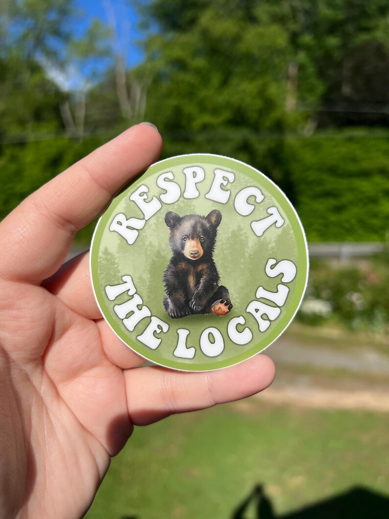 May include: A round sticker with a green background and white text that reads "Respect the Locals". A black bear cub is sitting in the centre of the sticker.