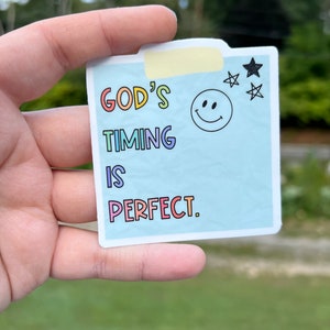 May include: A blue sticker with the text "GOD'S TIMING IS PERFECT." in rainbow colors. A smiley face and stars are in the top right corner.