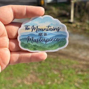 May include: A sticker with a cloud-shaped border featuring a watercolor mountain scene in shades of blue and green. The text "The Mountains are His Masterpiece" is written across the image in a script font.