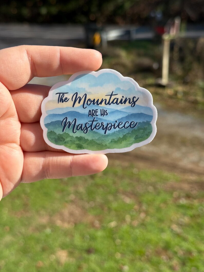 May include: A sticker with a cloud-shaped border featuring a watercolor mountain scene in shades of blue and green. The text "The Mountains are His Masterpiece" is written across the image in a script font.