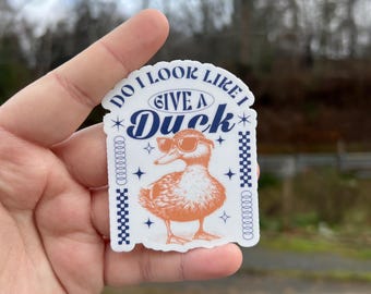 May include: A white sticker with blue and orange text and a cartoon duck wearing sunglasses. The text reads "Do I look like I give a Duck".