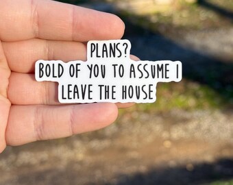 May include: A white sticker with black text that reads "PLANS? BOLD OF YOU TO ASSUME I LEAVE THE HOUSE". The sticker is held in a hand against a blurred outdoor background.