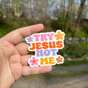 May include: A white sticker with the text "Try Jesus Not Me" in colorful, retro-style lettering. The sticker is decorated with pink, orange, and purple flowers.