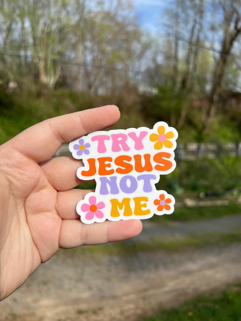 May include: A white sticker with the text "Try Jesus Not Me" in colorful, retro-style lettering. The sticker is decorated with pink, orange, and purple flowers.