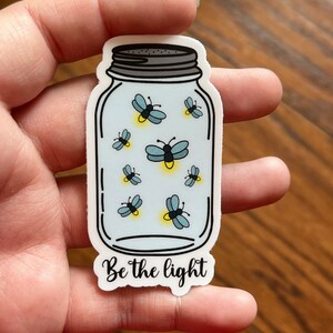 May include: A white sticker with a blue mason jar filled with glowing fireflies. The text "Be the light" is printed below the jar.