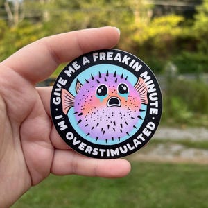 May include: A round sticker with a black background and a blue and turquoise border. The sticker features a cartoon pufferfish with pink, purple, and orange colors. The pufferfish has large, teary eyes and an open mouth. The text on the sticker reads "Give me a freakin minute. I'm overstimulated."