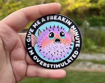 May include: A round sticker with a black background and a blue and turquoise border. The sticker features a cartoon pufferfish with pink, purple, and orange colors. The pufferfish has large, teary eyes and an open mouth. The text on the sticker reads "Give me a freakin minute. I'm overstimulated."