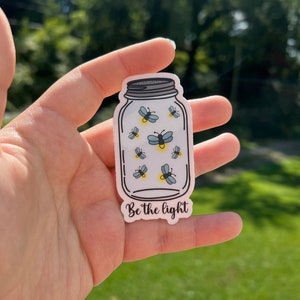 May include: A white sticker with a drawing of a mason jar filled with fireflies. The text "Be the light" is printed below the jar.