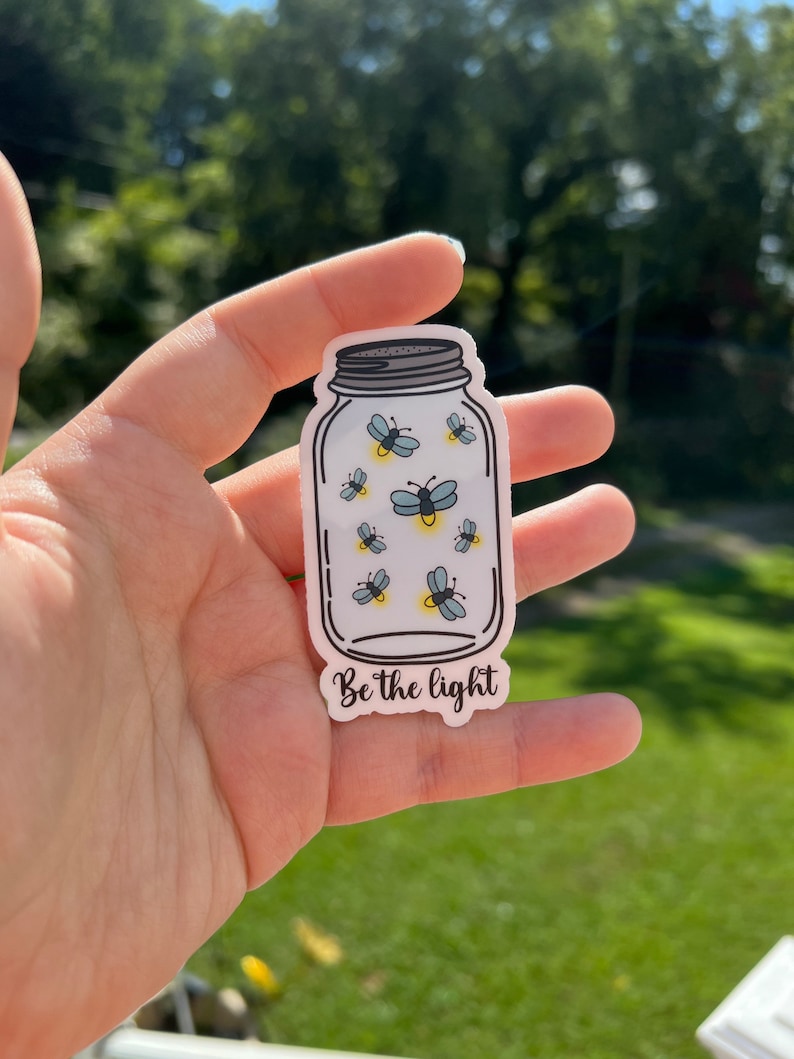 May include: A white sticker with a drawing of a mason jar filled with fireflies. The text "Be the light" is printed below the jar.
