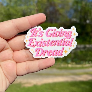 May include: Pink and white sticker with the text "It's Giving Existential Dread" in a retro font. The sticker has gold glitter accents.