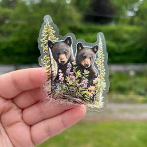 May include: A clear sticker with two black bear cubs peeking out from behind a cluster of green foliage and pink flowers.