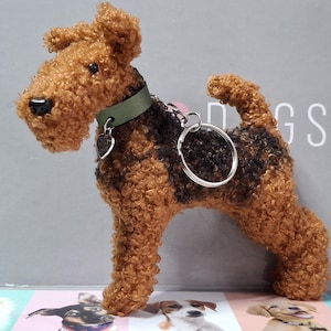 Op de afbeelding: Een bruine en zwarte pluche honden sleutelhanger met een groene halsband en een zilveren hartje. De hond staat op alle vier poten en heeft een krullend vacht.