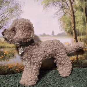 Peut inclure: Un chien en peluche fait à la main, avec une fourrure bouclée brun clair. Le chien porte un collier vert avec une étiquette en forme de cœur qui dit "With Love". L'arrière-plan présente un paysage peint avec des arbres et de l'eau.