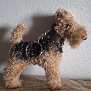 Op de afbeelding: Een sleutelhanger van een Airedale Terrier hond van pluche. De hond heeft een bruine en zwarte krullende vachttextuur, een bruine halsband en een zilveren sleutelring. De hond staat op een beige oppervlak.