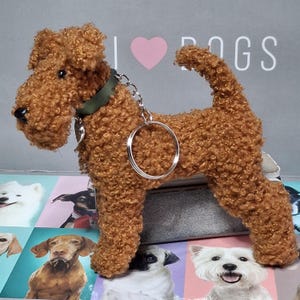 May include: A brown, fluffy, plush toy dog with a green collar and a silver key chain attached. The toy dog is standing on a surface with a background of various dog breeds.