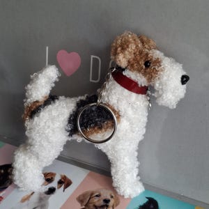 Puede incluir: Un llavero de perro de peluche hecho a mano en blanco, marrón y negro. El perro tiene un collar rojo y un llavero plateado. El fondo es una superficie gris claro con un corazón rosa y la letra "D".