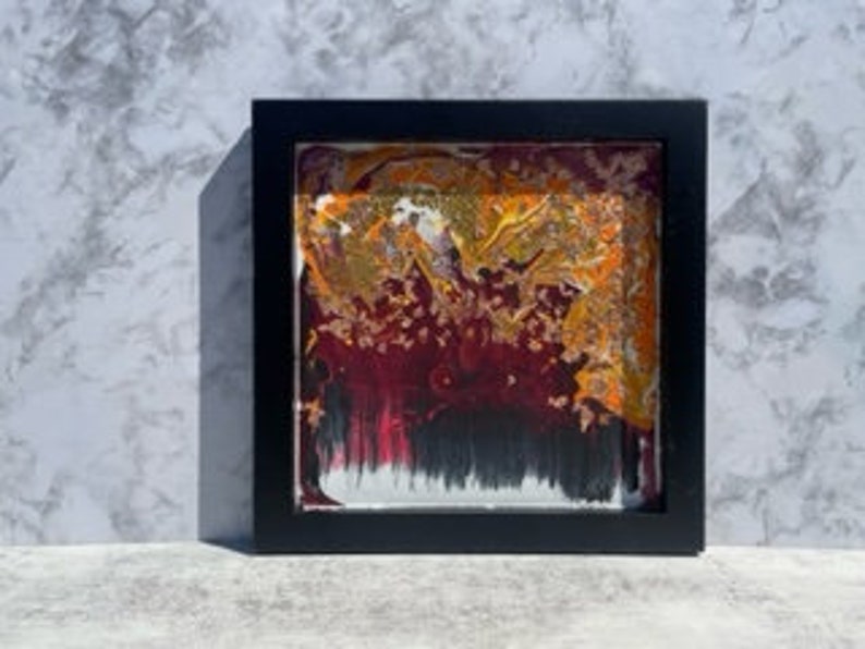 May include: Abstract art painting in a black frame. The painting features a swirling mix of red, orange, yellow, and black paint.