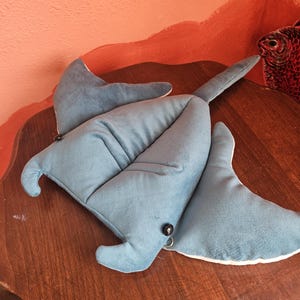 May include: A blue plush stingray toy with a white underside, black button eyes, and a long tail. The toy is lying on a wooden surface. The background features a painted wall with a fish.