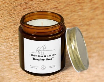 May include: A brown glass jar candle with a white label featuring a horse illustration and the text "Barn time is not like 'Regular time'". The lid is off to the side. The candle is on a brown surface.