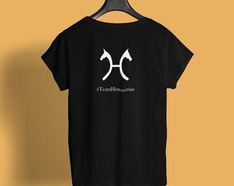 May include: Black t-shirt with a white graphic of two stylized horse heads facing each other. Below the graphic is the text "#TeamHanoverian". The shirt is hanging on a black hanger against a yellow background.