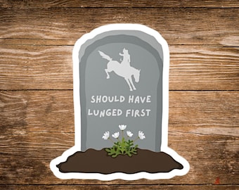 May include: A sticker featuring a gray tombstone with the text "SHOULD HAVE LUNGED FIRST". A white silhouette of a person riding a horse is on the tombstone. White flowers and green foliage are at the base of the tombstone.
