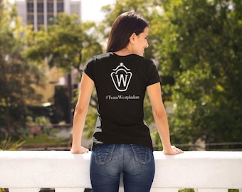 May include: Black t-shirt with a white graphic and the text "#TeamWestphalian". The shirt is worn by a person in blue jeans. The background features green trees and a white wall.