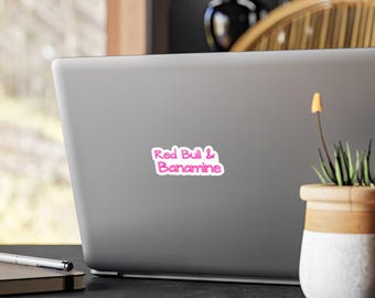 May include: A rectangular sticker with a white background and a thin pink border, featuring the text Red Bull & Banamine written in a stylized, bright pink cursive font. The sticker is applied to the center of a closed silver laptop lid.