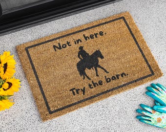 May include: A rectangular doormat with a black border and the text "Not in here. Try the barn." A black silhouette of a person riding a horse is in the center. Sunflowers and gardening gloves are in the image.