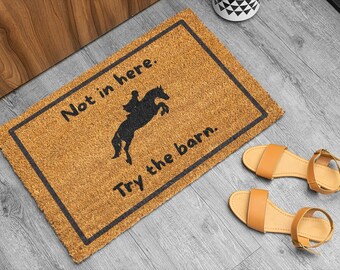 May include: A rectangular doormat with a black border and the text "Not in here. Try the barn." features a black silhouette of a horse and rider. Two pairs of tan sandals are next to the doormat.