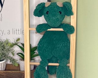 May include: A green crocheted dragon plush toy with brown horns and black eyes is sitting on a wooden ladder. The dragon is holding onto the ladder with its front paws. The text "@FoxyMommaCrochet" is written on the wooden floor below the ladder.