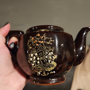 May include: A brown teapot with a gold and white floral design. The teapot has a spout and a handle.