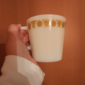 May include: A white mug with a gold floral pattern around the rim. The mug has a handle and a cylindrical shape. The design features stylized flowers and leaves. The mug is held by a person.