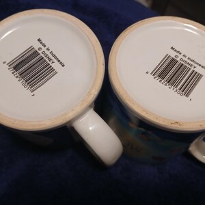 Op de afbeelding: Twee witte keramische mokken met handvatten. De bodem van elke mok toont een barcode, de tekst "Made in Indonesia" en het Disney copyright symbool. Kleurrijk ontwerp.