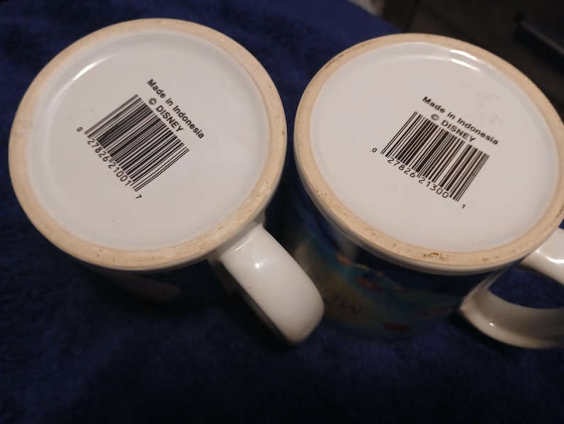 Op de afbeelding: Twee witte keramische mokken met handvatten. De bodem van elke mok toont een barcode, de tekst "Made in Indonesia" en het Disney copyright symbool. Kleurrijk ontwerp.