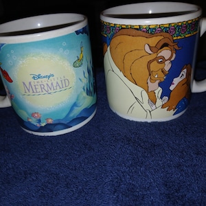 Op de afbeelding: Twee witte keramische mokken. E&eacute;n toont Disney's De Kleine Zeemeermin met een onderwater sc&egrave;ne. De andere mok toont het Beest en Belle van Belle en het Beest. Beide mokken hebben witte handvatten.
