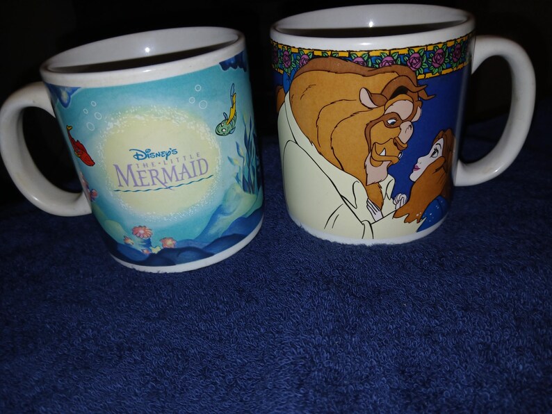 Op de afbeelding: Twee witte keramische mokken. E&eacute;n toont Disney's De Kleine Zeemeermin met een onderwater sc&egrave;ne. De andere mok toont het Beest en Belle van Belle en het Beest. Beide mokken hebben witte handvatten.