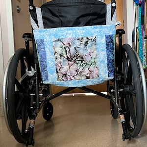 Peut inclure: Un fauteuil roulant noir avec un sac en tissu bleu et blanc attaché à l'arrière. Le sac a une fenêtre avec un motif de papillon.