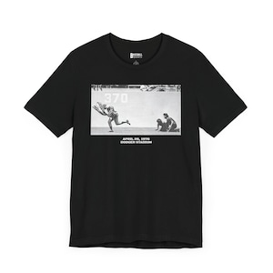 May include: Black t-shirt with a vintage photograph of a baseball player running with the number 370 on the wall behind him. The text below the image reads "April 26, 1976 Dodger Stadium".