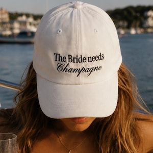 May include: A white baseball cap featuring the embroidered black text The Bride needs Champagne. The cap has a structured crown and a curved brim. The fabric is a solid white cotton material with matching white stitching and a button on top.
