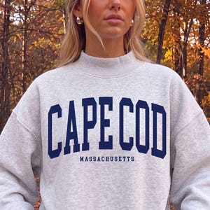 May include: A light gray heathered crewneck sweatshirt with a mock neck collar. The front features the dark blue text CAPE COD in a bold collegiate font, with the word MASSACHUSETTS printed in a smaller, matching dark blue font below it.