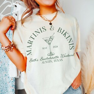 May include: A white t-shirt with a green graphic of a martini glass and the text "Martinis & Bikinis" in a circle. The text "Beth's Bachelorette Weekend Austin, Texas" is below the circle. The text "May 2024" is inside the circle.