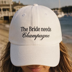 Może przedstawiać: Biała czapka z daszkiem z czarnym haftowanym napisem The Bride needs Champagne. Czapka posiada usztywnianą koronę, zakrzywiony daszek oraz przeszywane oczka na górnych panelach. Haft wykonany jest przy użyciu mieszanki czcionek szeryfowych i skryptowych.