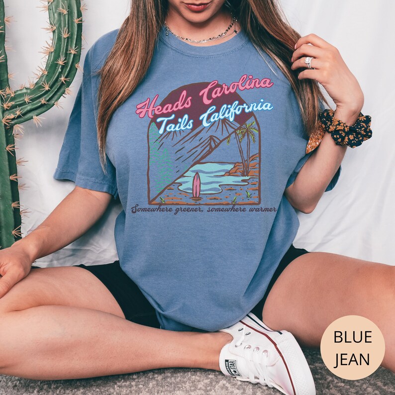 May include: A blue jean t-shirt with a graphic of a beach scene and the text "Heads Carolina Tails California" and "Somewhere greener, somewhere warmer".