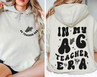 May include: A light beige hoodie with a black graphic print on the front that says "Ag Teacher Era" with a corn cob design. The back of the hoodie has a black graphic print that says "In My Ag Teacher Era" with a corn cob and cow design.