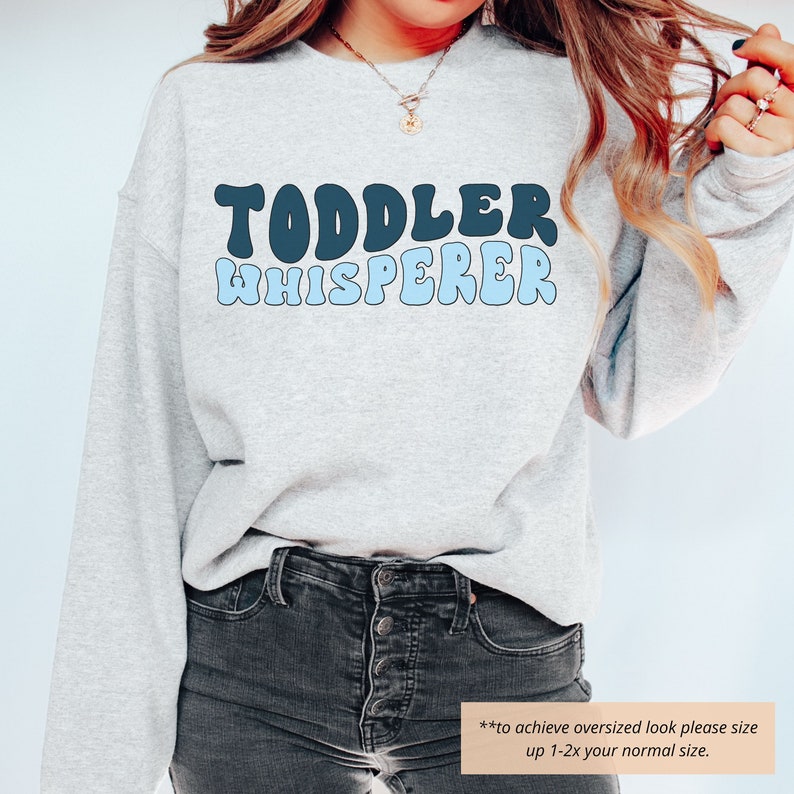 Puede incluir: Una sudadera gris claro con el texto "TODDLER WHISPERER" en letras retro azules y negras. El texto est&aacute; en un degradado de azul y negro, con "TODDLER" en negro y "WHISPERER" en azul. La sudadera la lleva puesta una persona, y el texto es visible en la parte delantera de la sudadera.