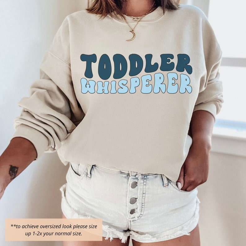 Puede incluir: Una sudadera de color beige claro con el texto "TODDLER WHISPERER" en letras azules y azul oscuro. El texto est&aacute; en una fuente retro y ondulada. La sudadera la lleva puesta una persona con las manos en los bolsillos. El texto "to achieve oversized look please size up 1-2x your normal size." est&aacute; en la parte inferior de la imagen.