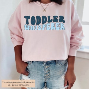 Puede incluir: Una sudadera rosa con el texto "TODDLER WHISPERER" en letras azules y azul claro. El texto est&aacute; en un estilo de fuente retro. La sudadera est&aacute; siendo usada por una persona. Para lograr un look de gran tama&ntilde;o, aumenta la talla de 1 a 2 veces tu talla normal.