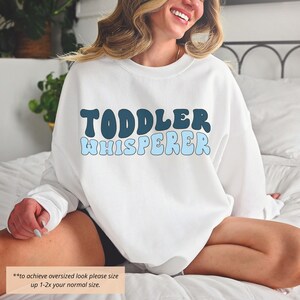 Puede incluir: Sudadera blanca con texto azul y azul oscuro que dice "Toddler Whisperer" en una fuente retro. El texto est&aacute; centrado en la sudadera. La modelo lleva la sudadera y est&aacute; sentada en una cama.