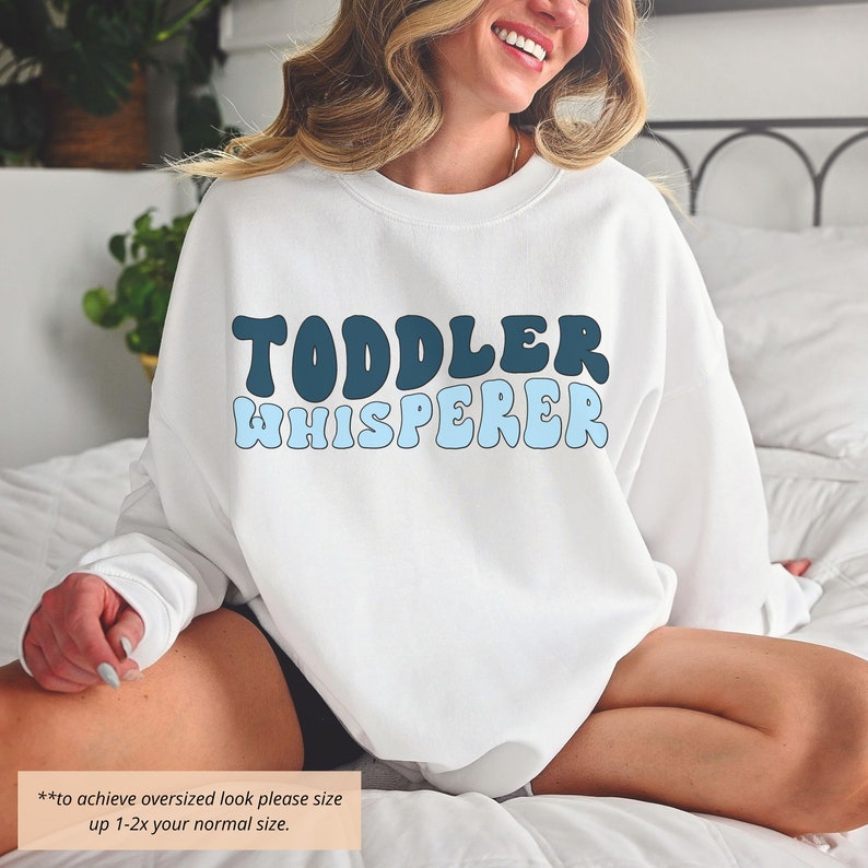 Puede incluir: Sudadera blanca con texto azul y azul oscuro que dice "Toddler Whisperer" en una fuente retro. El texto est&aacute; centrado en la sudadera. La modelo lleva la sudadera y est&aacute; sentada en una cama.